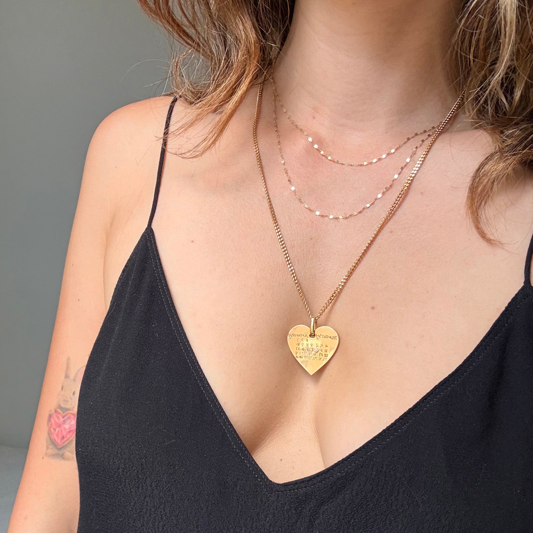 A close-up of a model wearing three layered necklaces: a gold heart-shaped pendant necklace with engraved text, a delicate 2-strand gold chain necklace.